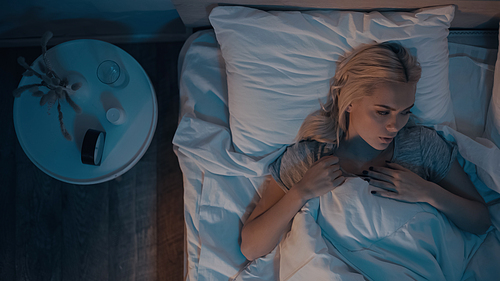 Top view of woman lying on bed near alarm clock, pills and water on bedside table