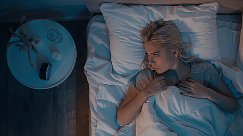 Top view of woman with insomnia looking at alarm clock near pills in bedroom