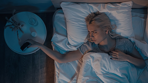 Top view of young woman with insomnia looking at alarm clock near pills and water on bedside table
