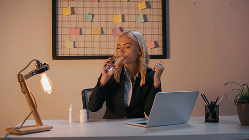 Businesswoman drinking water near laptop, pills and nasal spray on table