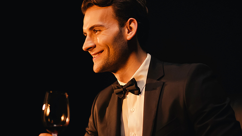 happy young man in suit holding glass of red wine isolated on black