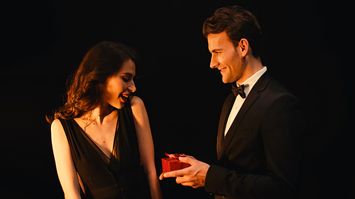 excited young woman receiving gift box from man in suit isolated on black