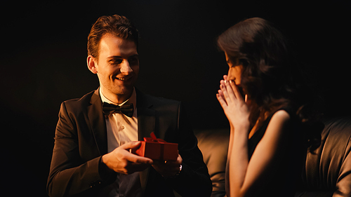 happy young man in suit with bow tie giving present to stunned woman isolated on black