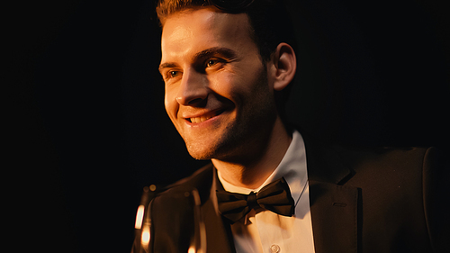 happy young man in suit with bow tie holding glass isolated on black