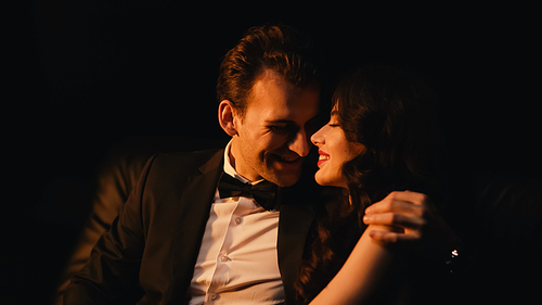 Smiling man in suit hugging and flirting with cheerful girlfriend isolated on black