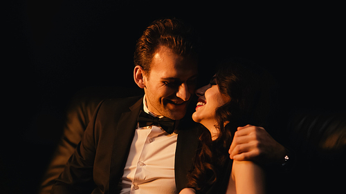 Smiling man in suit hugging and flirting with joyful girlfriend isolated on black