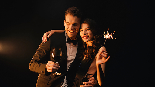 woman with sparkler hugging happy man holding glass of red wine on black