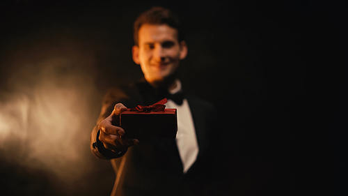happy and blurred man in suit showing red gift box on black