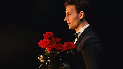 Side view of elegant young man holding red roses isolated on black