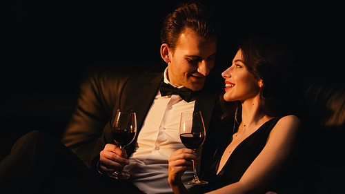 Happy couple holding glasses of red wine while looking at each other isolated on black
