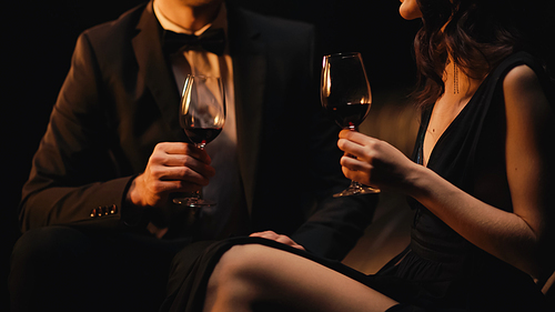 cropped view of couple holding glasses of red wine while sitting on sofa isolated on black