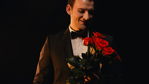 happy young man looking at red roses and smiling isolated on black