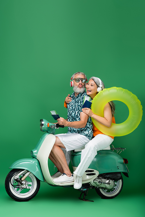 multiethnic middle aged couple in wireless headphones holding passports with air tickets and riding motor scooter on green