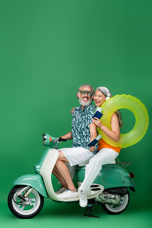 happy and multiethnic mature couple in wireless headphones holding passports with air tickets and riding motor scooter on green