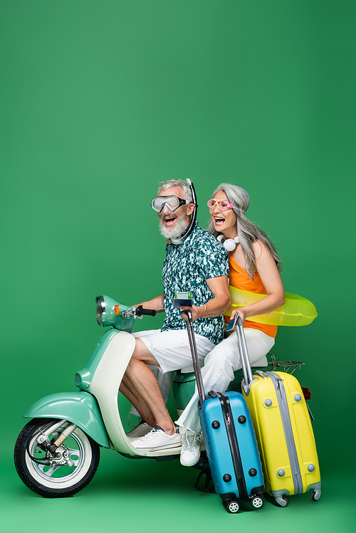 excited and multiethnic middle aged couple in goggles holding passports and luggage while riding motor scooter on green