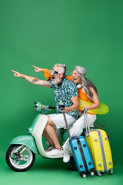 amazed and multiethnic middle aged couple in goggles pointing with fingers and holding passports while riding motor scooter on green