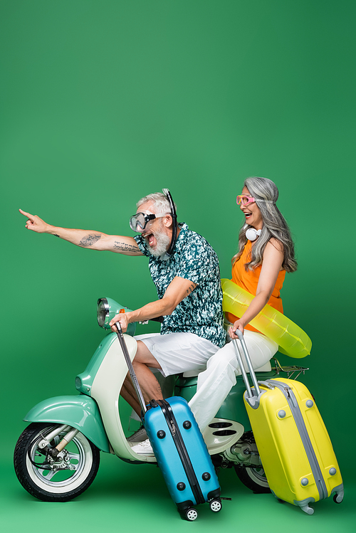 amazed and middle aged man in goggles pointing with finger near asian wife with luggage while riding motor scooter on green