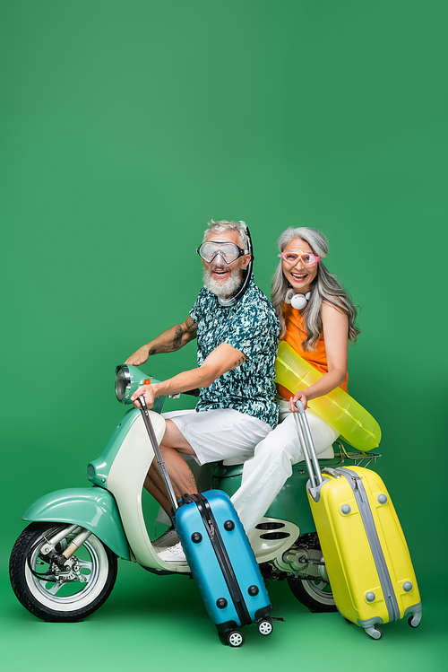 multiethnic and happy middle aged couple in goggles holding luggage while riding moped on green