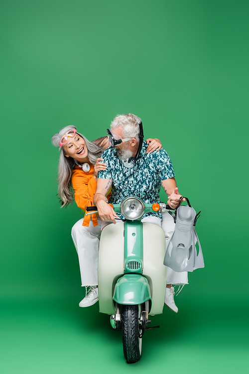 cheerful middle aged asian woman looking at husband in goggles holding flippers while riding motor scooter on green