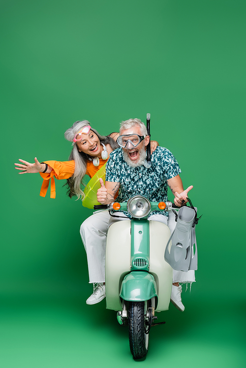 cheerful middle aged asian woman looking at husband in goggles holding flippers while showing thumbs up and riding moped on green