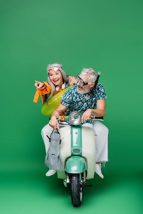 cheerful middle aged asian woman pointing with finger near husband holding flippers while riding motor scooter on green