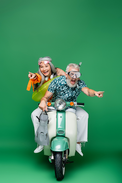 cheerful and multiethnic middle aged couple in goggles pointing with fingers while riding moped on green