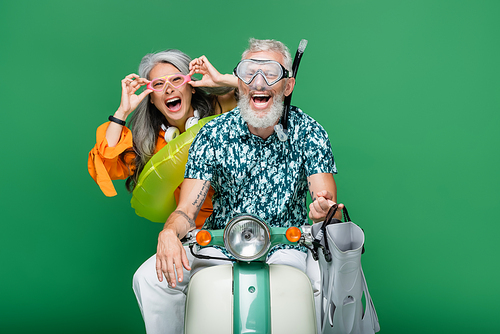multiethnic and excited middle aged couple in goggles riding moped isolated on green