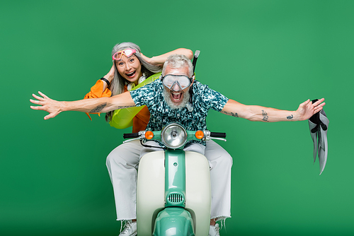cheerful middle aged man with outstretched hands holding flippers near asian wife in goggles riding moped on green