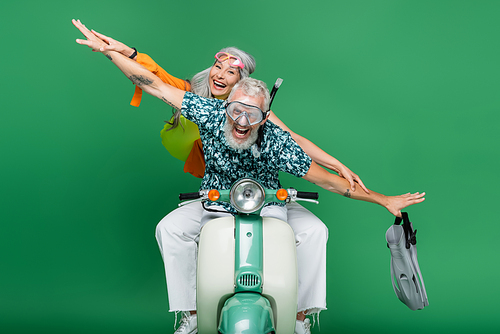 multiethnic and cheerful middle aged couple with outstretched hands riding moped on green