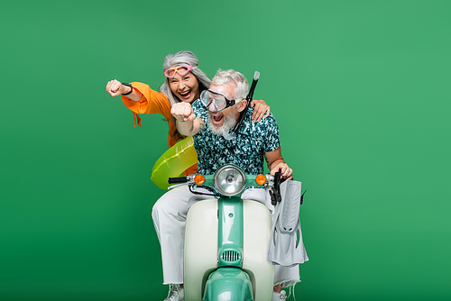 multiethnic and happy middle aged couple in goggles with outstretched hands riding moped on green