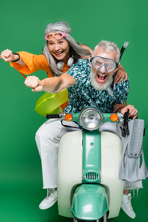 positive and multiethnic middle aged couple in goggles with outstretched hands riding motor scooter on green