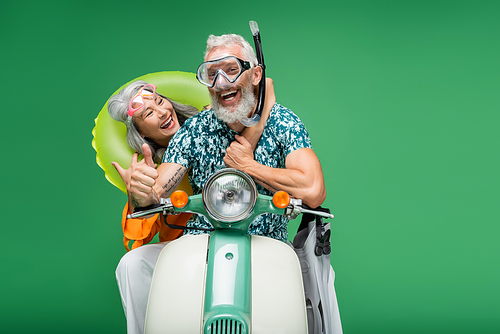 cheerful middle aged asian woman hugging husband in goggles showing thumb up while riding motor scooter isolated on green