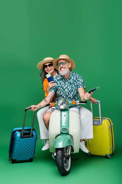 cheerful middle aged and multiethnic couple in sun hats holding passports and luggage while riding moped on green