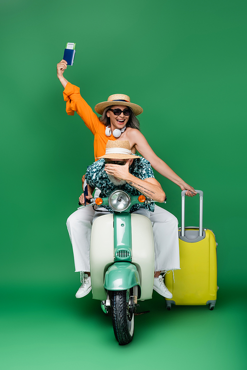cheerful middle aged asian woman in sun hat holding passport near husband covering eyes while riding motor scooter on green