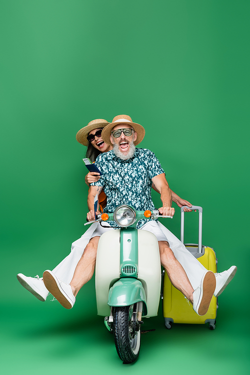 interracial and positive middle aged couple in sun hats holding passports with air tickets while riding moped on green