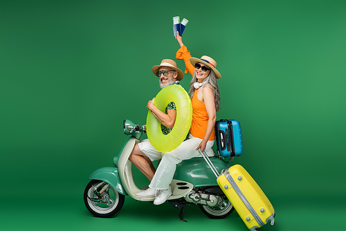 cheerful middle aged asian woman in sun hat holding passports near husband holding swim ring while riding moped on green