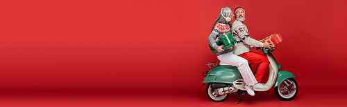 cheerful middle aged and asian woman in ear muffs holding christmas presents while riding moped with husband on red, banner