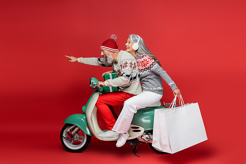cheerful middle aged asian woman holding shopping bags while riding moped with husband pointing with finger on red
