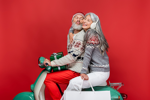 middle aged man with christmas presents pouting lips near asian wife while riding motor scooter on red