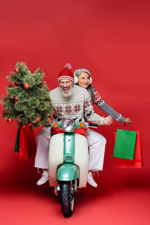 happy middle aged man holding christmas tree near asian wife while riding moped on red