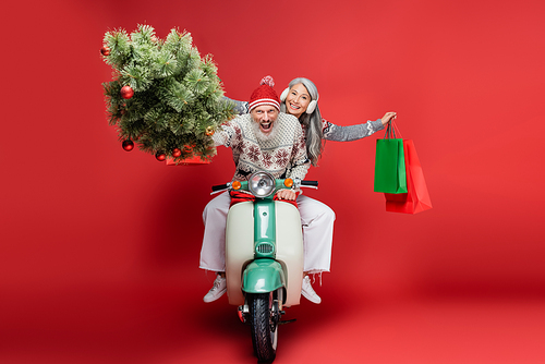 excited middle aged man holding christmas tree near asian wife while riding moped on red