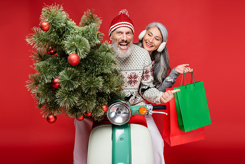 cheerful middle aged man holding christmas tree near asian wife while riding moped on red