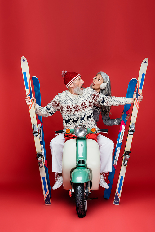 surprised and interracial middle aged couple in sweaters holding skis while riding motor scooter on red