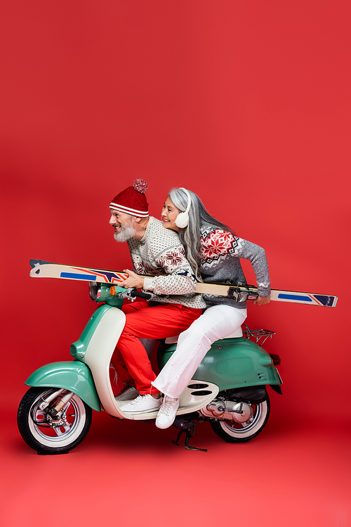 side view of happy interracial and middle aged couple in sweaters holding skis while riding moped on red