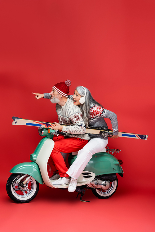 surprised and interracial middle aged woman in sweater pointing with finger near husband holding skis while riding moped on red