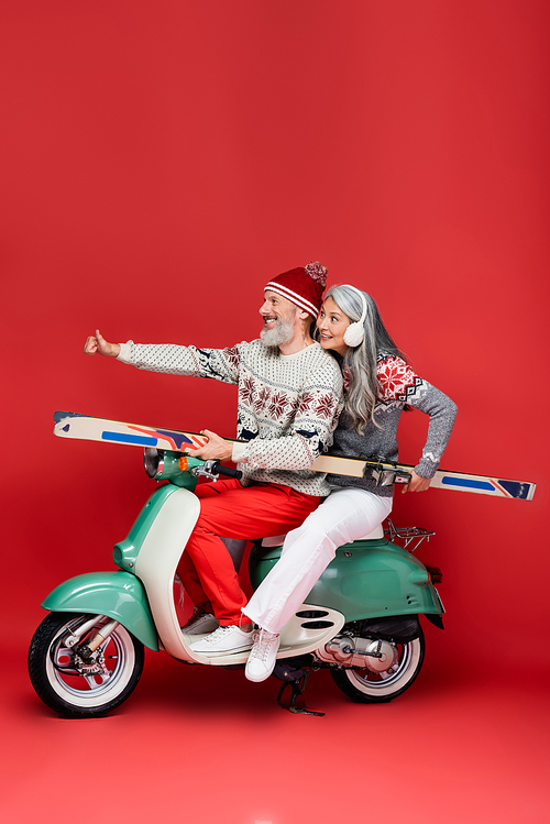 smiling interracial and middle aged woman in sweater holding skis while husband showing thumb up and riding moped on red