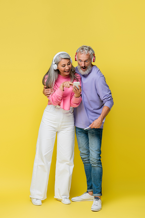 full length of happy interracial and middle aged woman in wireless headphones pointing at smartphone near surprised husband on yellow