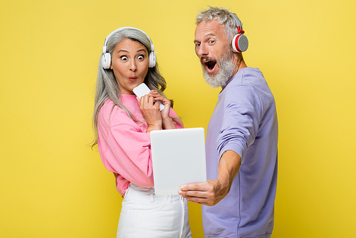 surprised middle aged and interracial couple in wireless headphones looking at digital tablet isolated on yellow