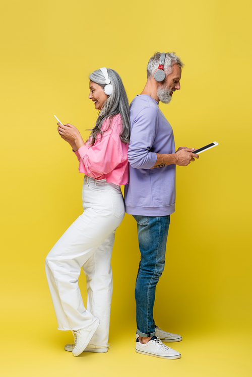 side view of cheerful middle aged and interracial couple in wireless headphones using gadgets while standing back to back on yellow