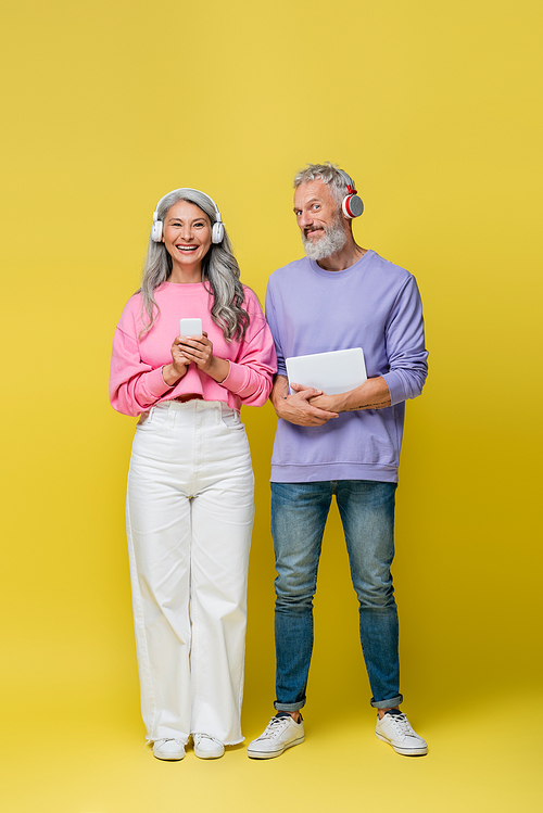 full length of multiethnic and smiling middle aged couple in wireless headphones holding gadgets on yellow